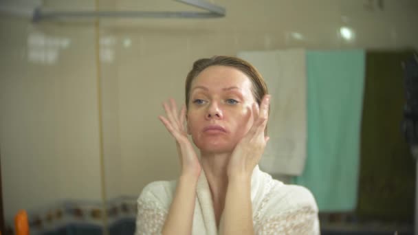 Femme en peignoir appliquer la crème sur le visage et le cou dans la salle de bain. 4k, au ralenti — Video