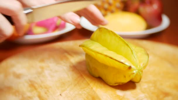 Exotiska frukter på bordet. 4k, kvinnliga händer skär en carambola med en kniv på en skärbräda — Stockvideo