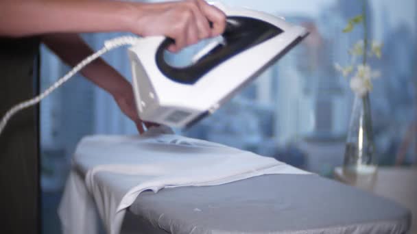 Рука женщины гладит белую рубашку утюгом перед панорамным окном с видом на небоскребы, 4k — стоковое видео