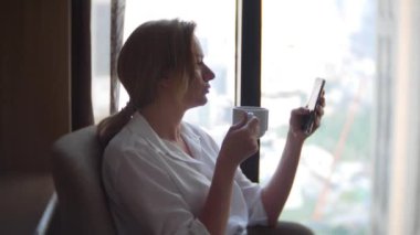 Kahve içme şehir manzaralı cam kenarında beyaz bir gömlek güzel kadın. ve onun smartphone 4 k kullanır