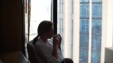 Güzel kadın pencerenin cityscape manzaralı kahve içme Beyaz gömlekli. 4k