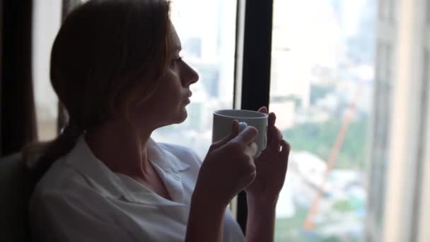 Beautiful woman in a white shirt drinking coffee by the window with cityscape view. 4k — Stock Video