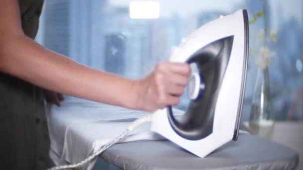 A womans hand strokes a white shirt with an iron in front of a panoramic window overlooking the skyscrapers, 4k — Stock Video