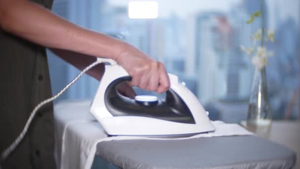 A womans hand strokes a white shirt with an iron in front of a panoramic window overlooking the skyscrapers, 4k — Stock Video