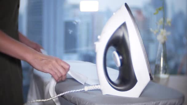 A womans hand strokes a white shirt with an iron in front of a panoramic window overlooking the skyscrapers, 4k — Stock Video