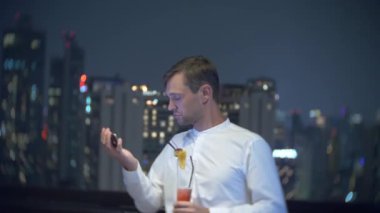 Genç, bir smartphone kullanarak ve bir bar kokteyl içme güzel adam geceleri gökdelenler bakan teras. 4k, arka plan bulanıklık.