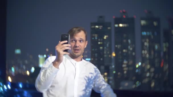 Νέοι, όμορφο άνδρα χρησιμοποιώντας ένα smartphone και πίνοντας ένα κοκτέιλ, σε ένα μπαρ, βεράντα με θέα τους ουρανοξύστες τη νύχτα. 4k, θάμπωμα του φόντου. — Αρχείο Βίντεο