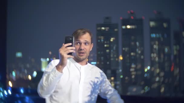 Giovane, bellissimo uomo che usa uno smartphone e beve un cocktail, su una terrazza bar con vista sui grattacieli di notte. 4k, sfocatura dello sfondo . — Video Stock