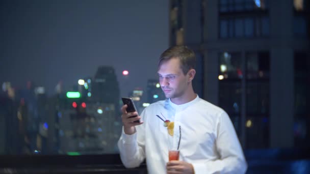 Giovane, bellissimo uomo che usa uno smartphone e beve un cocktail, su una terrazza bar con vista sui grattacieli di notte. 4k, sfocatura dello sfondo . — Video Stock