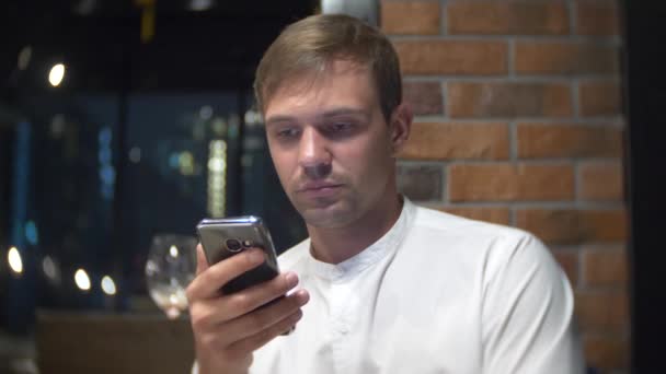 Um homem irritado a verificar um telemóvel à noite num restaurante. 4k, borrão de fundo — Vídeo de Stock