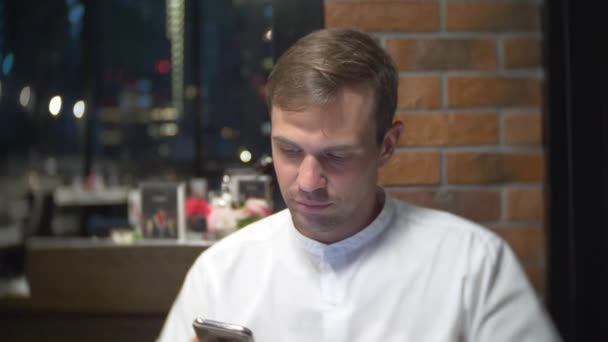 Een man drinkt koffie, een mobiele telefoon te controleren bij nacht in een restaurant. Hij kijkt naar het polshorloge. 4k, achtergrond vervagen — Stockvideo