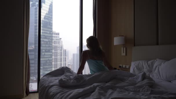 Женщина просыпается от сна и сидит на кровати на заднем плане окна с видом на небоскребы, медленное движение. 4k — стоковое видео