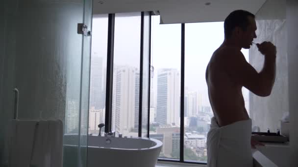 Young man, brushing teeth with a toothbrush in the bathroom with a panoramic window. view from the window to the skyscrapers. 4k, slow motion — Stock Video