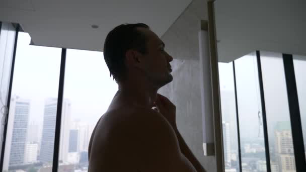 Красивый мужчина наносит бритву в ванной комнате с видом из окна на небоскребы. 4k, slow motion — стоковое видео