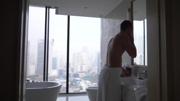 Un hombre guapo aplicando una navaja de afeitar en el baño con vista desde la ventana a los rascacielos. 4k, cámara lenta — Vídeos de Stock