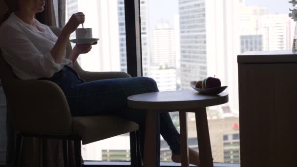 Gyönyörű nő és kávét iszik az ablak kilátás nyílik a városra, a felhőkarcolók. 4k, lassú mozgás. — Stock videók