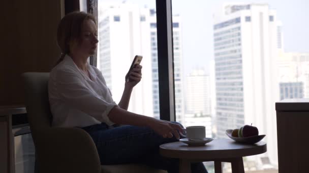 Krásná žena, pití kávy a pomocí její telefon u okna s výhledem na město, mrakodrapy. 4k, pomalý pohyb. — Stock video