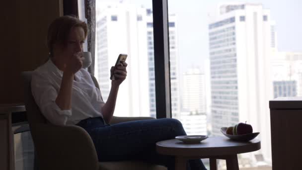 Hermosa mujer tomando café y usando su teléfono en la ventana con vistas a la ciudad, rascacielos. 4k, cámara lenta . — Vídeo de stock