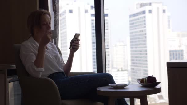 Schöne Frau Kaffee trinkt und ihr Telefon am Fenster mit Blick auf die Stadt, Wolkenkratzer. 4k, Zeitlupe. — Stockvideo