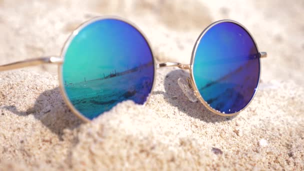 Gafas de sol acostadas en la arena de la playa. Las gafas de sol reflejan el mar, el sol, el cielo, la playa. 4k, cámara lenta — Vídeo de stock