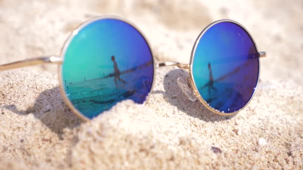 Gafas de sol acostadas en la arena de la playa. Las gafas de sol reflejan el mar, el sol, el cielo y la mujer caminando por la playa. 4k, cámara lenta — Vídeo de stock