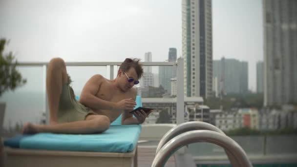 Un giovane uomo con un tablet in piscina sul tetto con vista sul mare. 4k. , sfocare lo sfondo — Video Stock