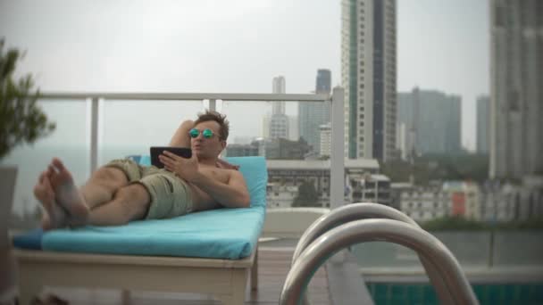Um jovem com um tablet na piscina no telhado com vista para o mar. 4K. , borrar o fundo — Vídeo de Stock
