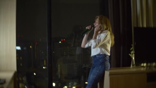 Een vrouw is bier drinken thuis naast een panoramisch venster met uitzicht op de wolkenkrabbers. nacht, achtergrond vervagen. 4k, slow-motion — Stockvideo