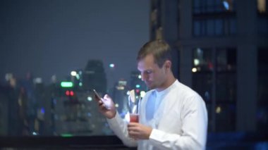 Genç, bir smartphone kullanarak ve bir bar kokteyl içme güzel adam geceleri gökdelenler bakan teras. 4k, arka plan bulanıklık.