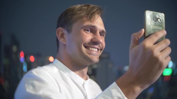 Joven y hermoso hombre usando un teléfono inteligente y bebiendo un cóctel, en una terraza del bar con vistas a los rascacielos por la noche. 4k, desenfoque de fondo . — Vídeos de Stock