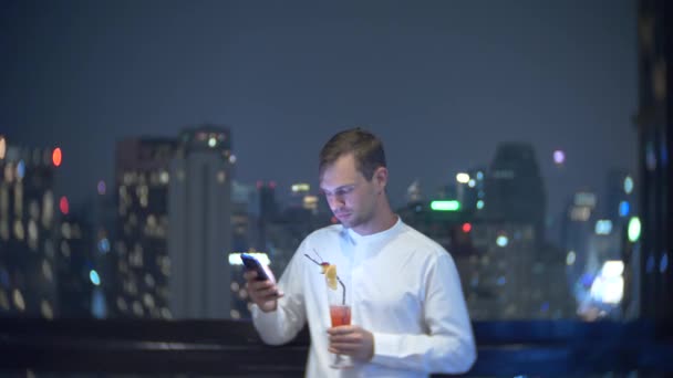 年轻, 漂亮的男子使用智能手机和喝鸡尾酒, 在酒吧露台俯瞰摩天大楼在夜间。4k. 背景模糊. — 图库视频影像
