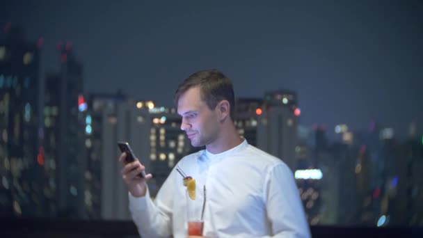 Giovane, bellissimo uomo che usa uno smartphone e beve un cocktail, su una terrazza bar con vista sui grattacieli di notte. 4k, sfocatura dello sfondo . — Video Stock