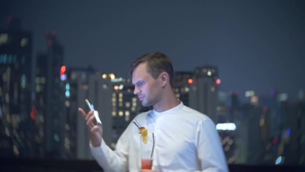 Giovane, bellissimo uomo che usa uno smartphone e beve un cocktail, su una terrazza bar con vista sui grattacieli di notte. 4k, sfocatura dello sfondo . — Video Stock