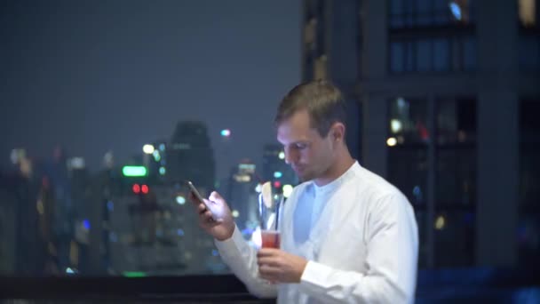 Junger, schöner Mann, der ein Smartphone benutzt und einen Cocktail trinkt, nachts auf einer Bar-Terrasse mit Blick auf die Wolkenkratzer. 4k, Hintergrund verschwommen. — Stockvideo