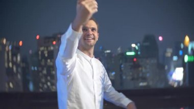 Genç, bir smartphone kullanarak ve bir bar kokteyl içme güzel adam geceleri gökdelenler bakan teras. 4k, arka plan bulanıklık.