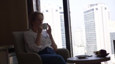 Kahve içme şehir manzaralı cam kenarında beyaz bir gömlek güzel kadın. ve onun smartphone 4 k kullanır