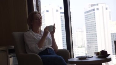 Kahve içme şehir manzaralı cam kenarında beyaz bir gömlek güzel kadın. ve onun smartphone 4 k kullanır