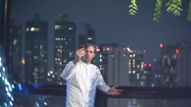 Νέοι, όμορφο άνδρα χρησιμοποιώντας ένα smartphone και πίνοντας ένα κοκτέιλ, σε ένα μπαρ, βεράντα με θέα τους ουρανοξύστες τη νύχτα. 4k, θάμπωμα του φόντου. — Αρχείο Βίντεο