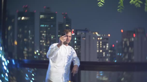 Giovane, bellissimo uomo che usa uno smartphone e beve un cocktail, su una terrazza bar con vista sui grattacieli di notte. 4k, sfocatura dello sfondo . — Video Stock