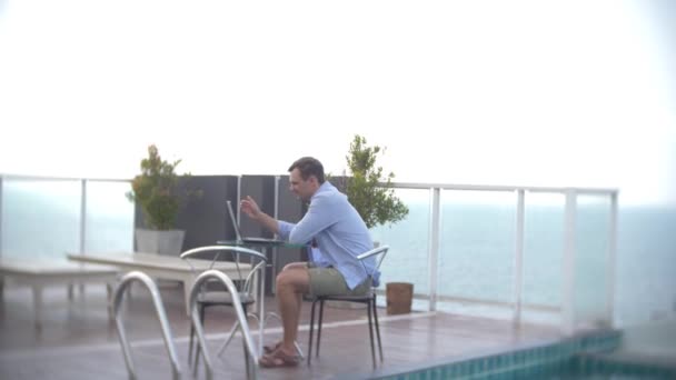 Empresario con portátil junto a la piscina en la azotea con vistas al mar. 4k. , difuminar el fondo — Vídeo de stock