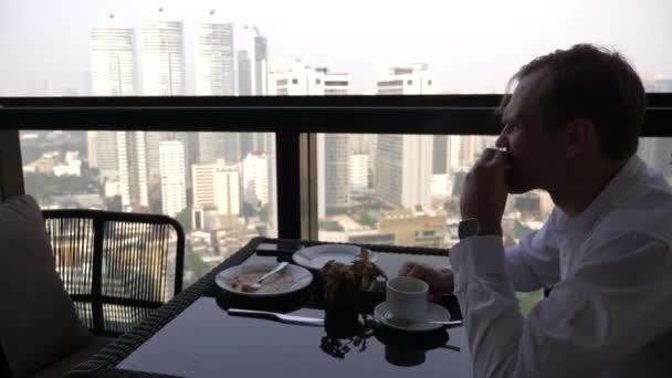 Młody biznesmen jest posiadanie śniadanie na tarasie restauracji z widokiem na wieżowce. 4k. — Wideo stockowe