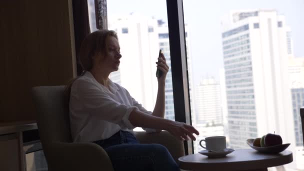 Красива жінка в білій сорочці п'є каву біля вікна з видом на місто. і використовує свій смартфон 4k — стокове відео