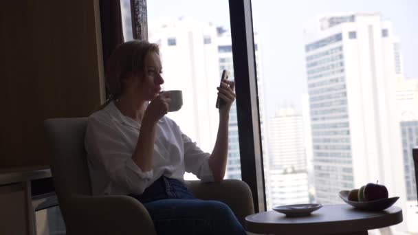 Mooie vrouw in een wit overhemd koffie drinken bij het raam met uitzicht op de stad. en gebruikt zijn smartphone 4k — Stockvideo