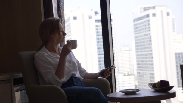 Красива жінка в білій сорочці п'є каву біля вікна з видом на місто. і використовує свій смартфон 4k — стокове відео