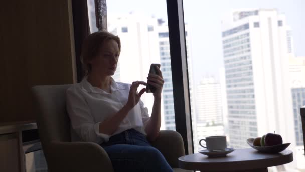 Hermosa mujer en una camisa blanca bebiendo café junto a la ventana con vistas a la ciudad. y utiliza su teléfono inteligente 4k — Vídeo de stock