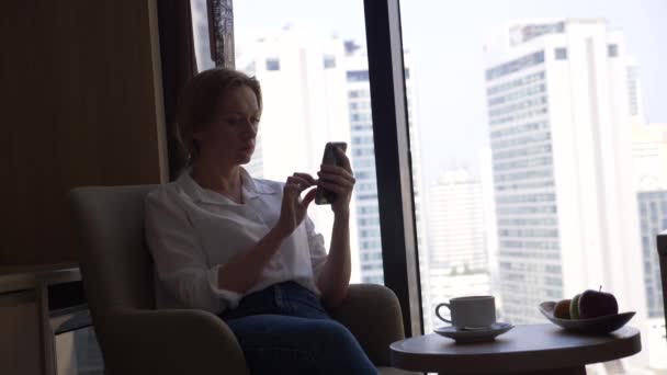 Hermosa mujer en una camisa blanca bebiendo café junto a la ventana con vistas a la ciudad. y utiliza su teléfono inteligente 4k — Vídeo de stock