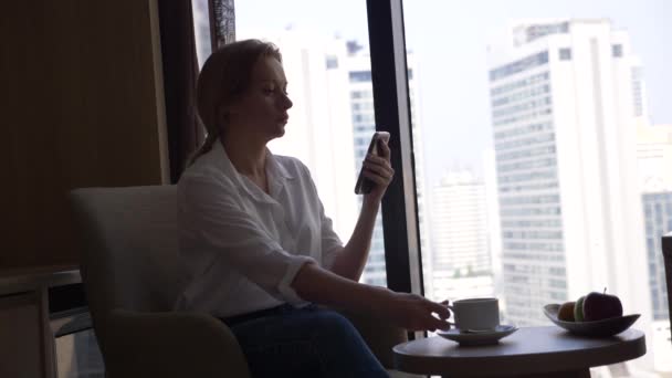 Hermosa mujer en una camisa blanca bebiendo café junto a la ventana con vistas a la ciudad. y utiliza su teléfono inteligente 4k — Vídeo de stock