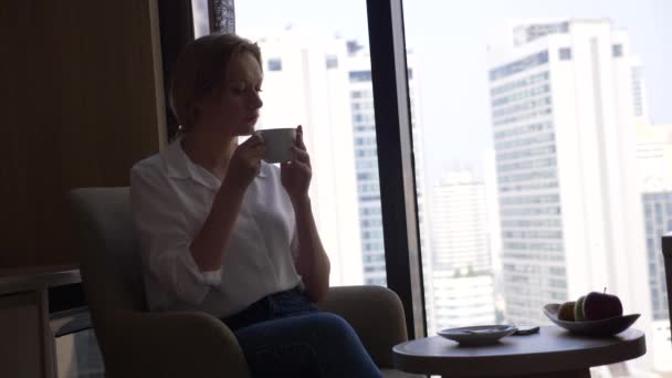 Красивая женщина в белой рубашке пьет кофе у окна с видом на город. и использует свой смартфон 4k — стоковое видео