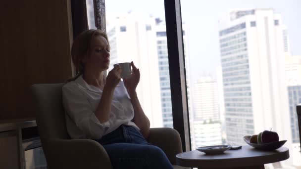 Piękna kobieta w białej koszuli, pije kawę przy oknie z widokiem na miasto. i używa jego smartfon 4k — Wideo stockowe