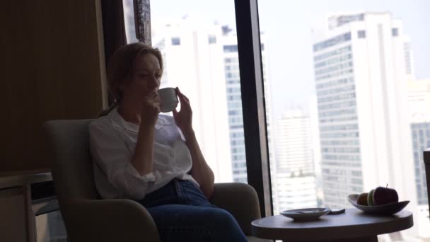 Όμορφη γυναίκα σε ένα λευκό πουκάμισο που πίνει καφέ από το παράθυρο με θέα στην πόλη. και χρήσεις του smartphone 4k — Αρχείο Βίντεο